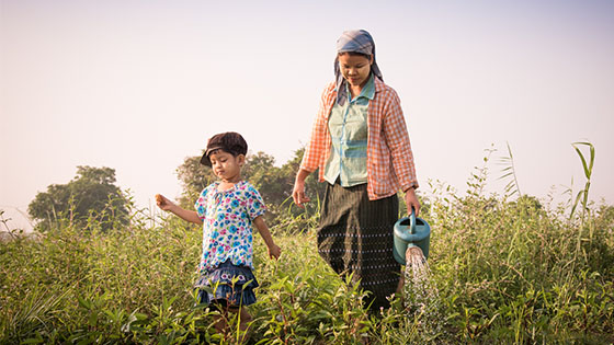 woman watering plants with a can while a small child walks in front of her on a grassy field.