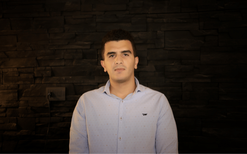 Hasan stands in front of the wall, wearing a blue button polo.