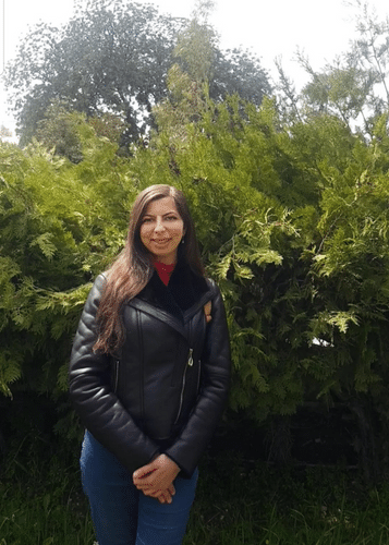 Riyam has long brown hair and is wearing a black leather jacket, standing in front of a hedge.