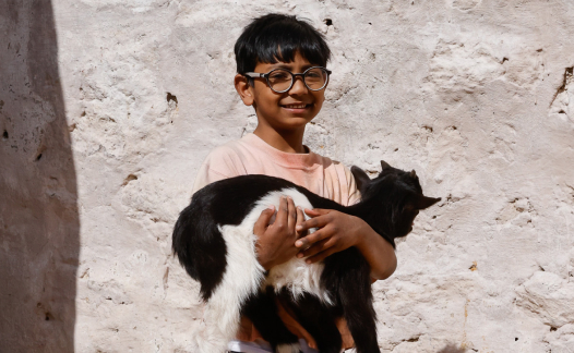 A boy wearing glasses, stands in front of a white wall, smiling and holding a goat.