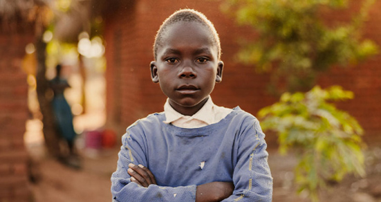 A young boy wearing a blue sweater and white collar stands with his arms crossed, looking directly at the camera, in an outdoor setting with blurred brick buildings and greenery.