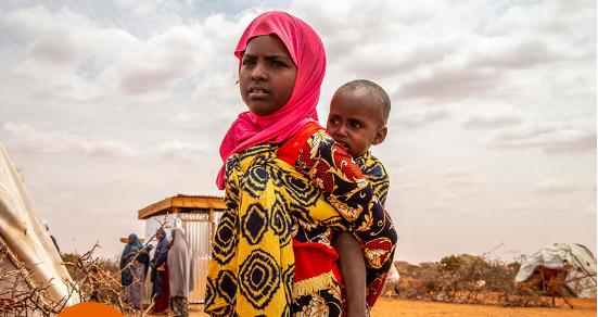 A young girl in a vibrant yellow and black patterned dress with a pink headscarf carries a small child on her back in a dry, arid landscape with scattered shelters.