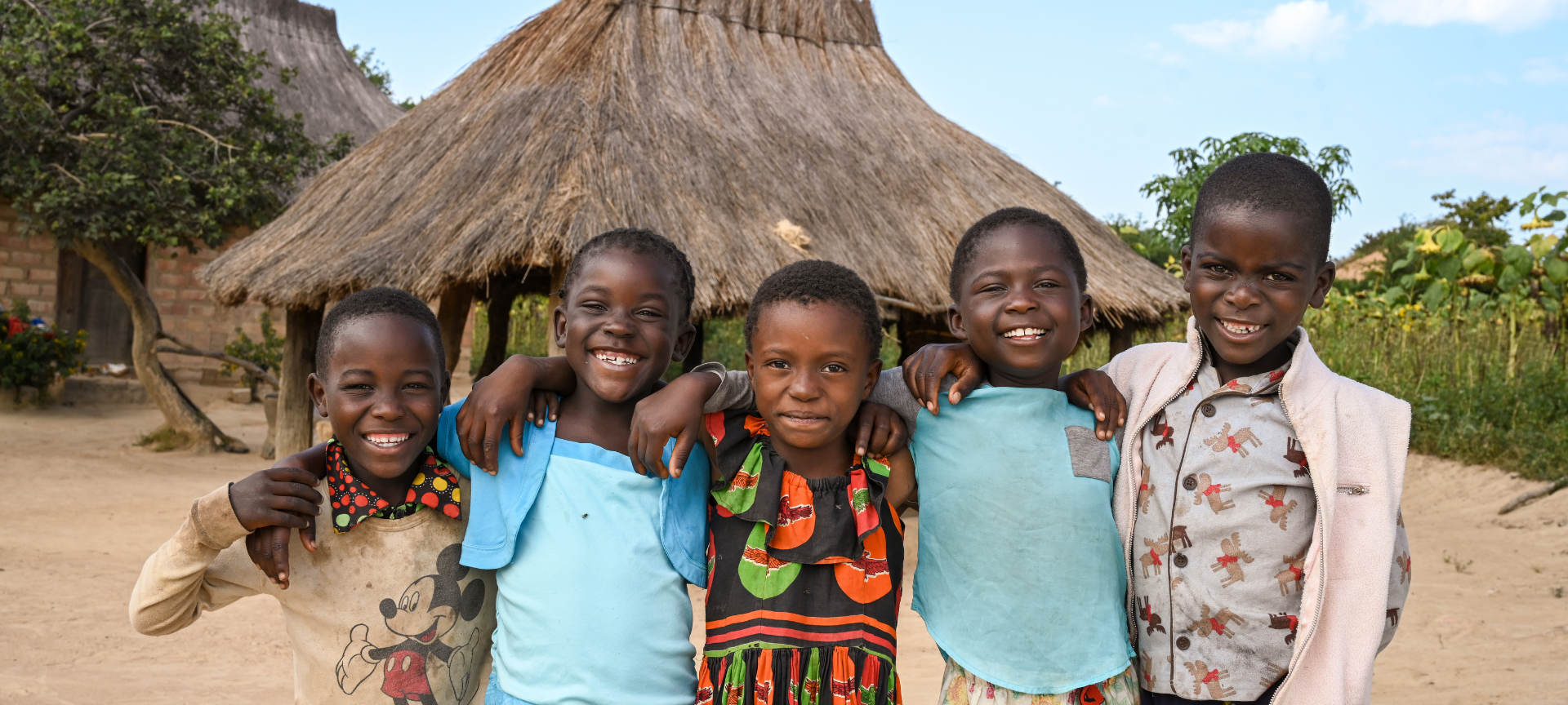 A group of f ive smiling children standing arm in arm in front of a hut, posing for a photo.