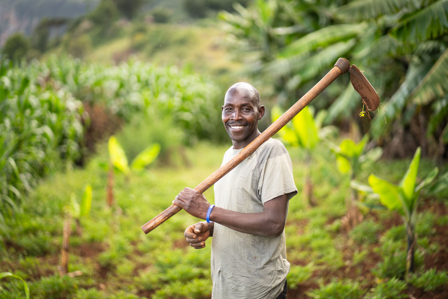 A Kenyan man carries a gardening hoe over his shoulder in his field, smiling at the camera. 