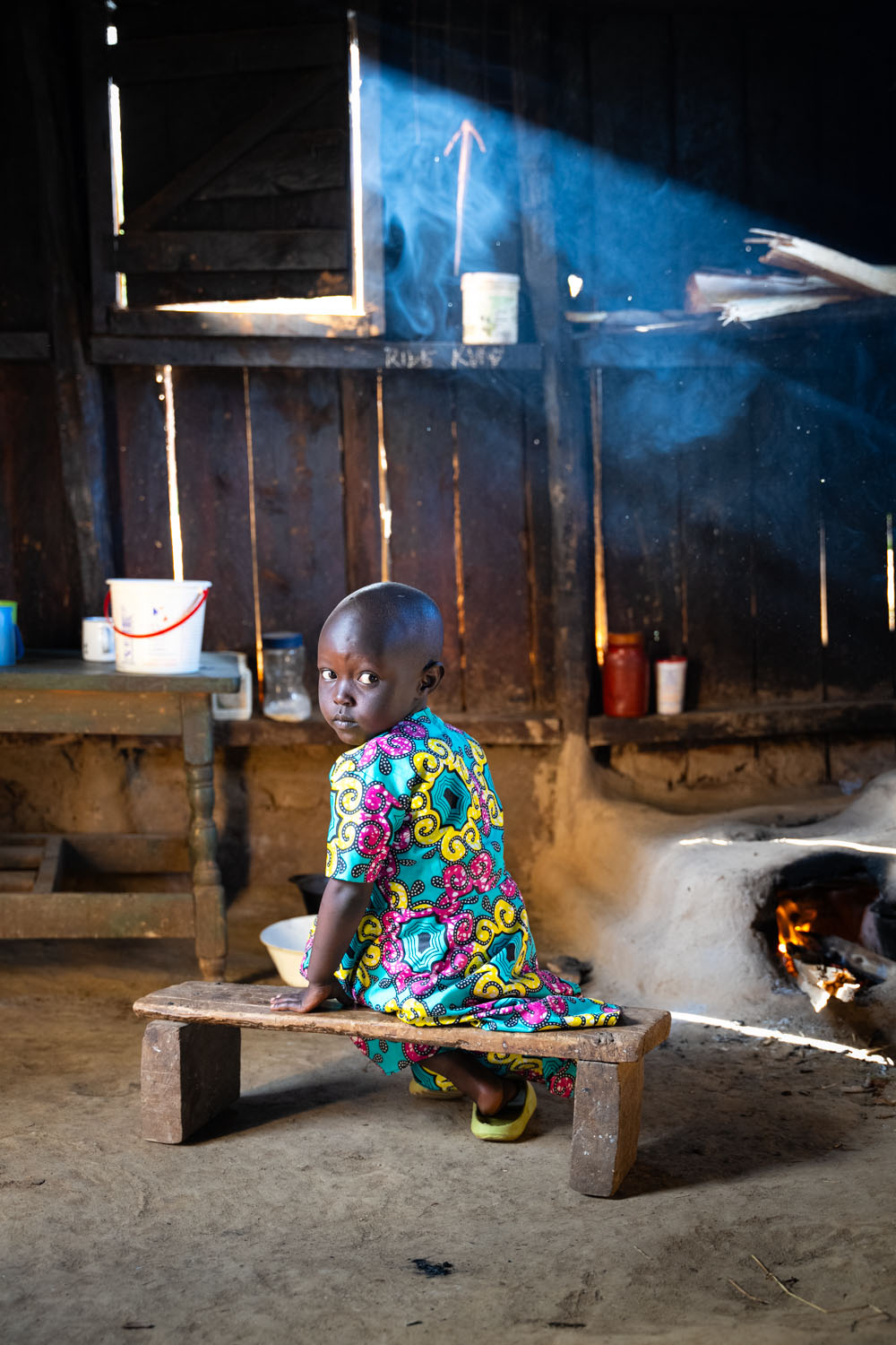 A young Kenyan girl sits on a small bench inside her house, looking over her shoulder, with a stove behind her. 