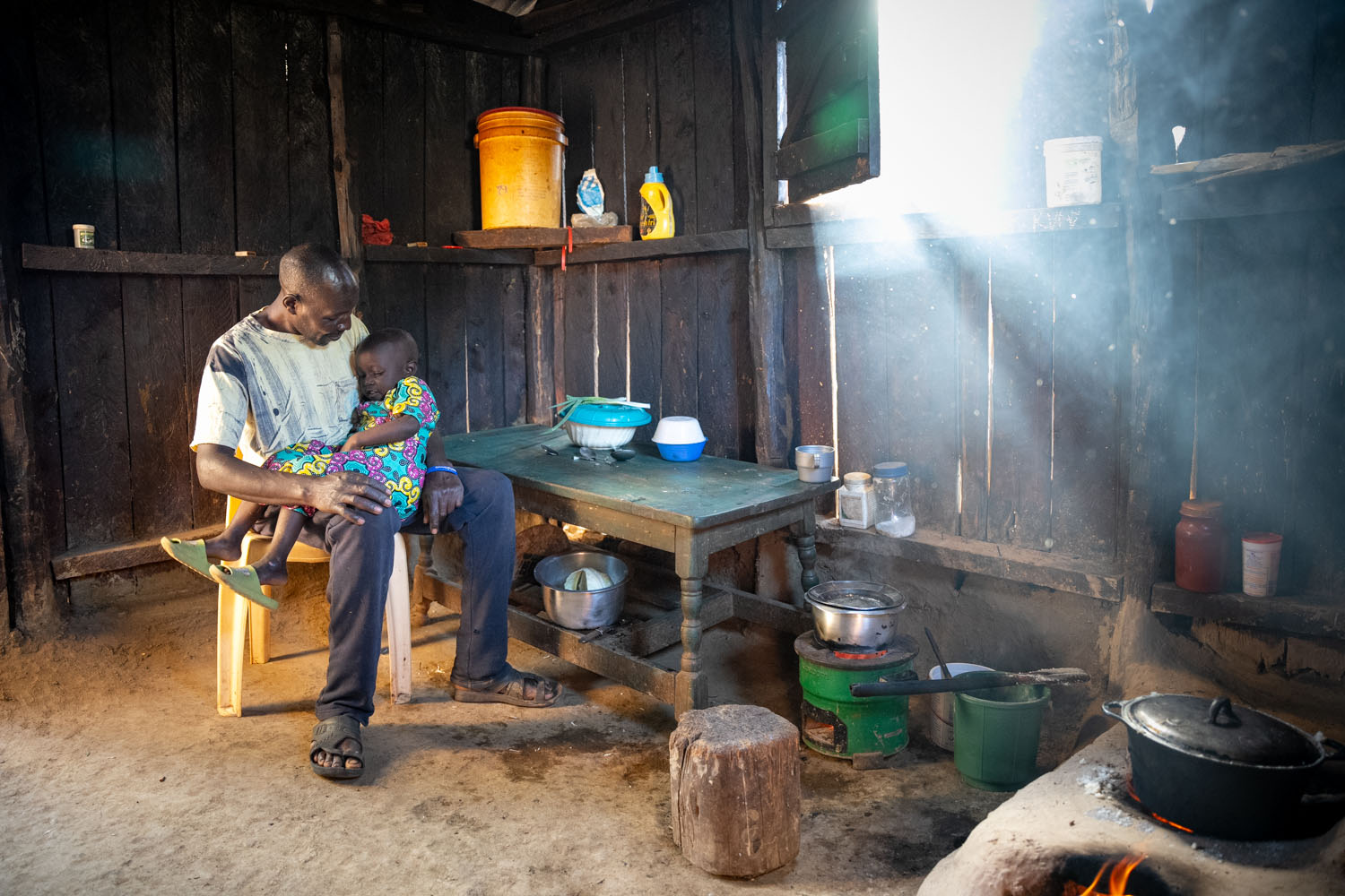 A Kenyan father holds his daughter in his lap, sitting in their home in front of their kitchen table. 
