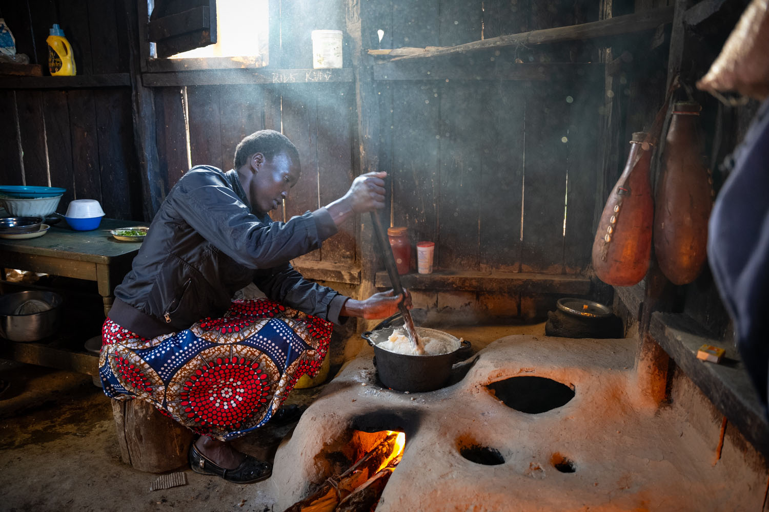 A Kenyan woman sits in front of her clay stove, stirring food in a pot over an open flame. 
