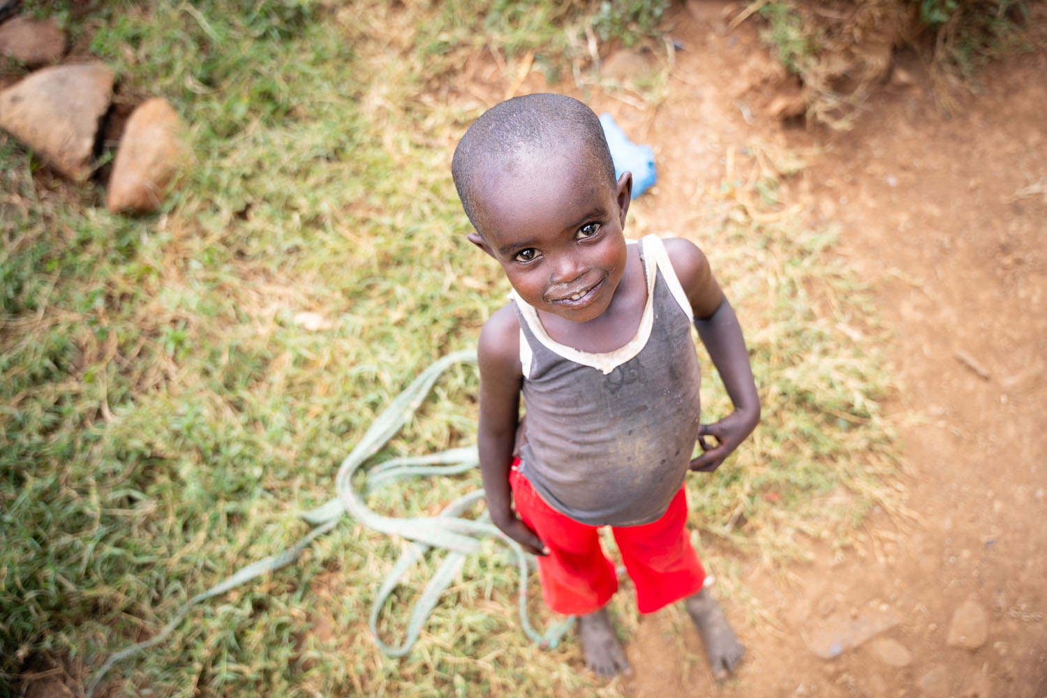 A Kenyan toddler boy wearing a gray tank top and red shorts smiles up at the camera. 