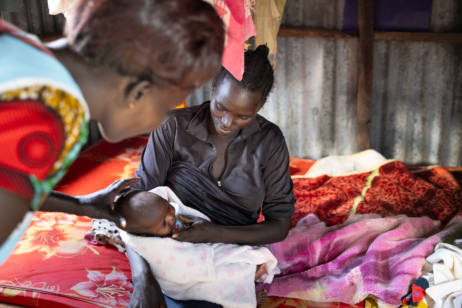 A Kenyan mother sits on her bed, holding her child, with a staff member leaning over and looking at the child. 