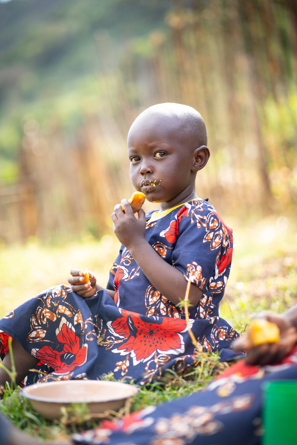 A young Kenyan girl sits on the ground outside her home, eating a sweet potato. 
