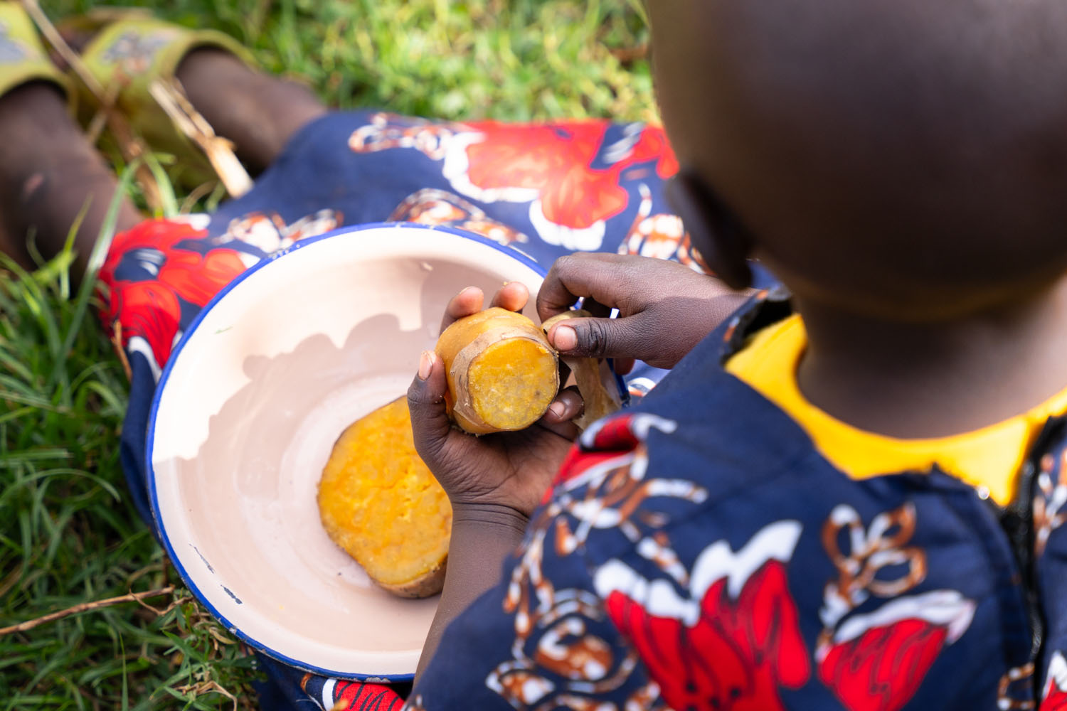 A child holds a sweet potato in her left hand with a plate in her lap, sitting in the grass. 