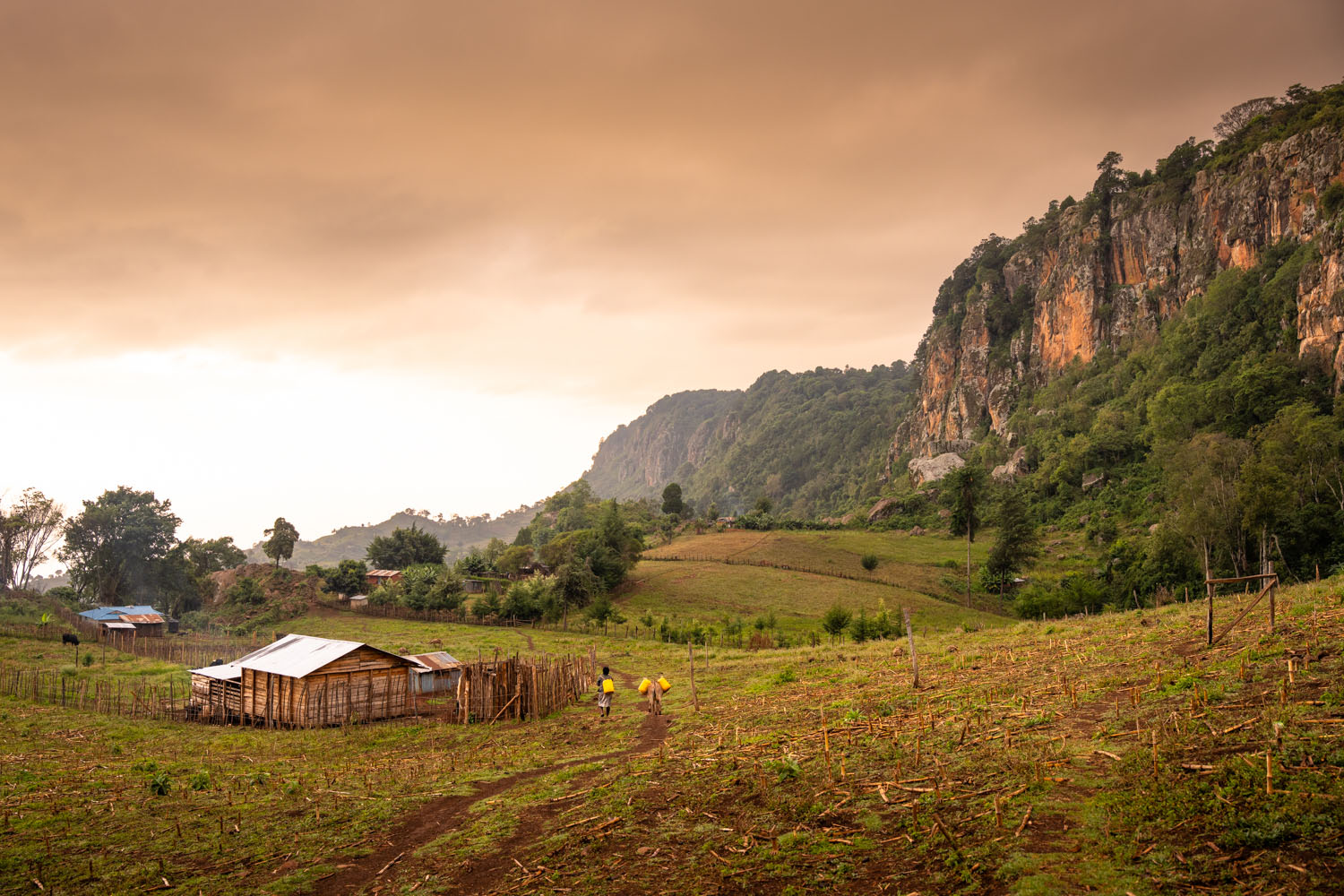 Two people walk towards a small house at the foot of cliffs in Rift Valley, Kenya. 