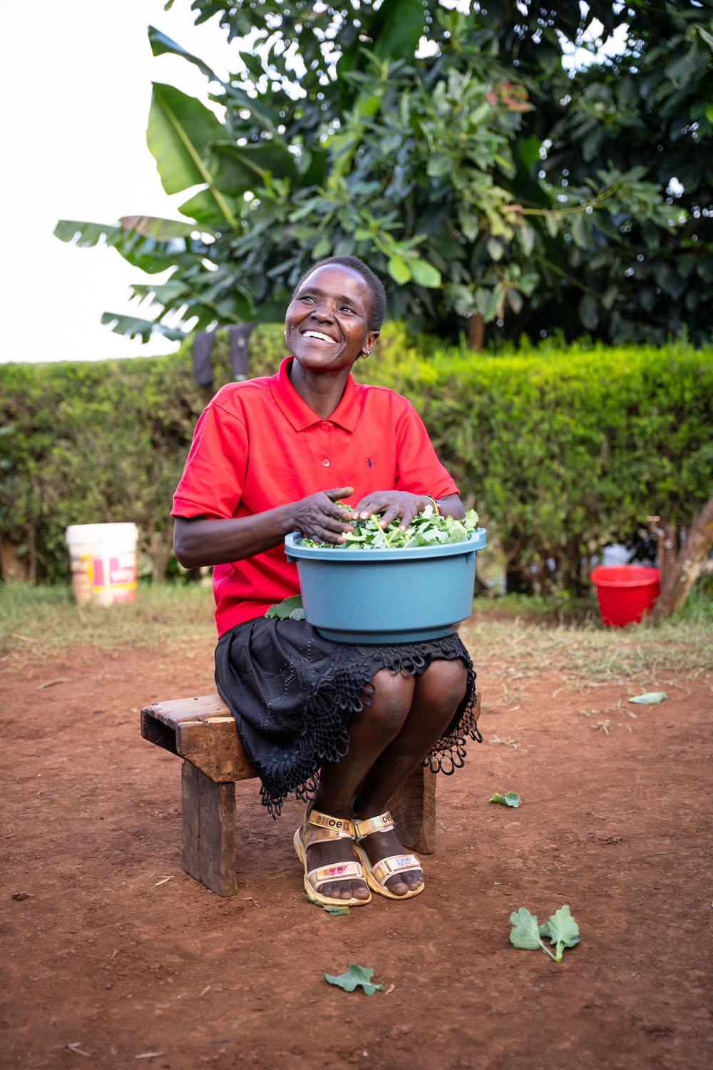 A Kenyan woman wearing a red polo shirt and black skirt sits on bench, sifting through vegetables in a basin. 