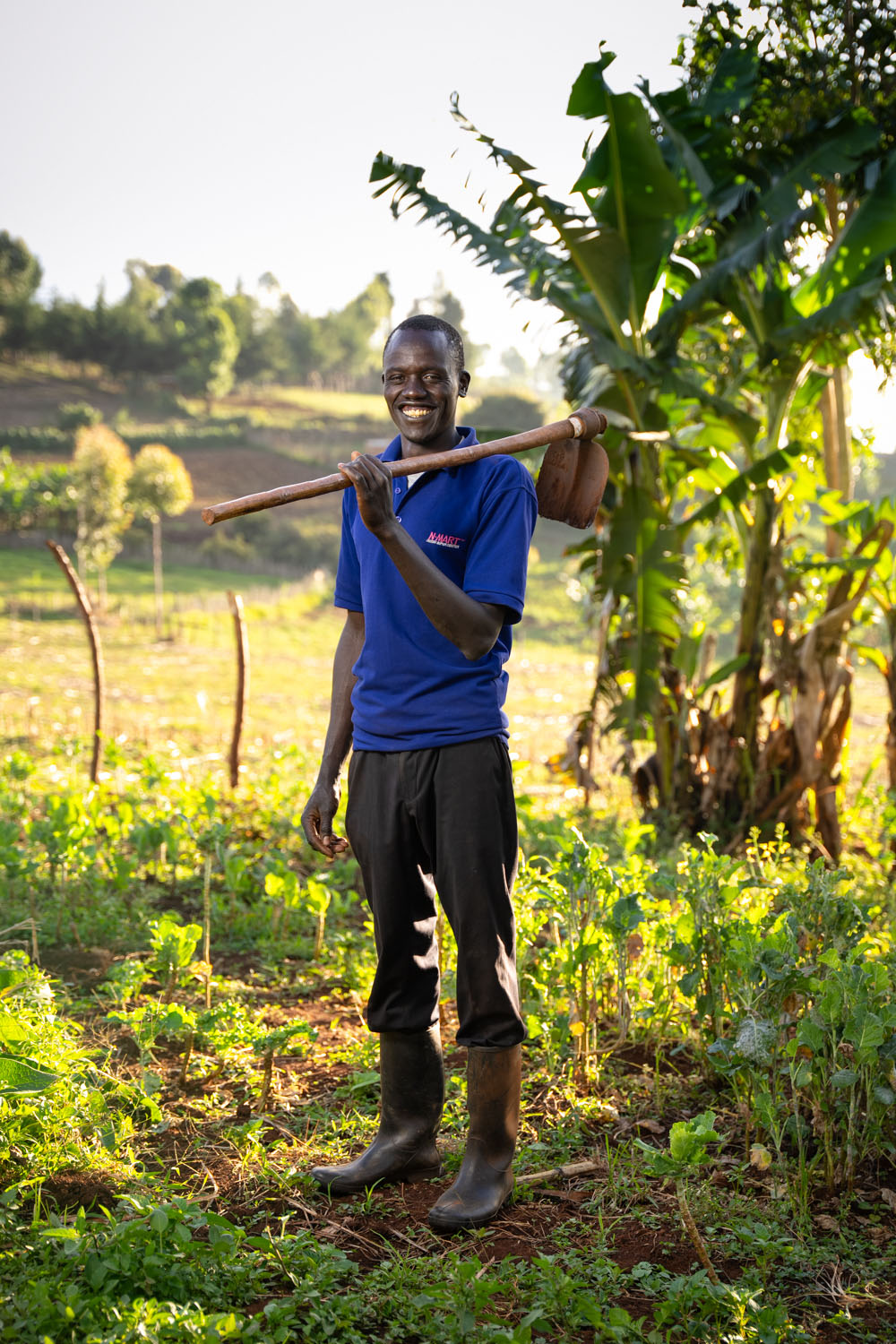 A Kenyan man stands in his field, holding a gardening hoe over his shoulder. 