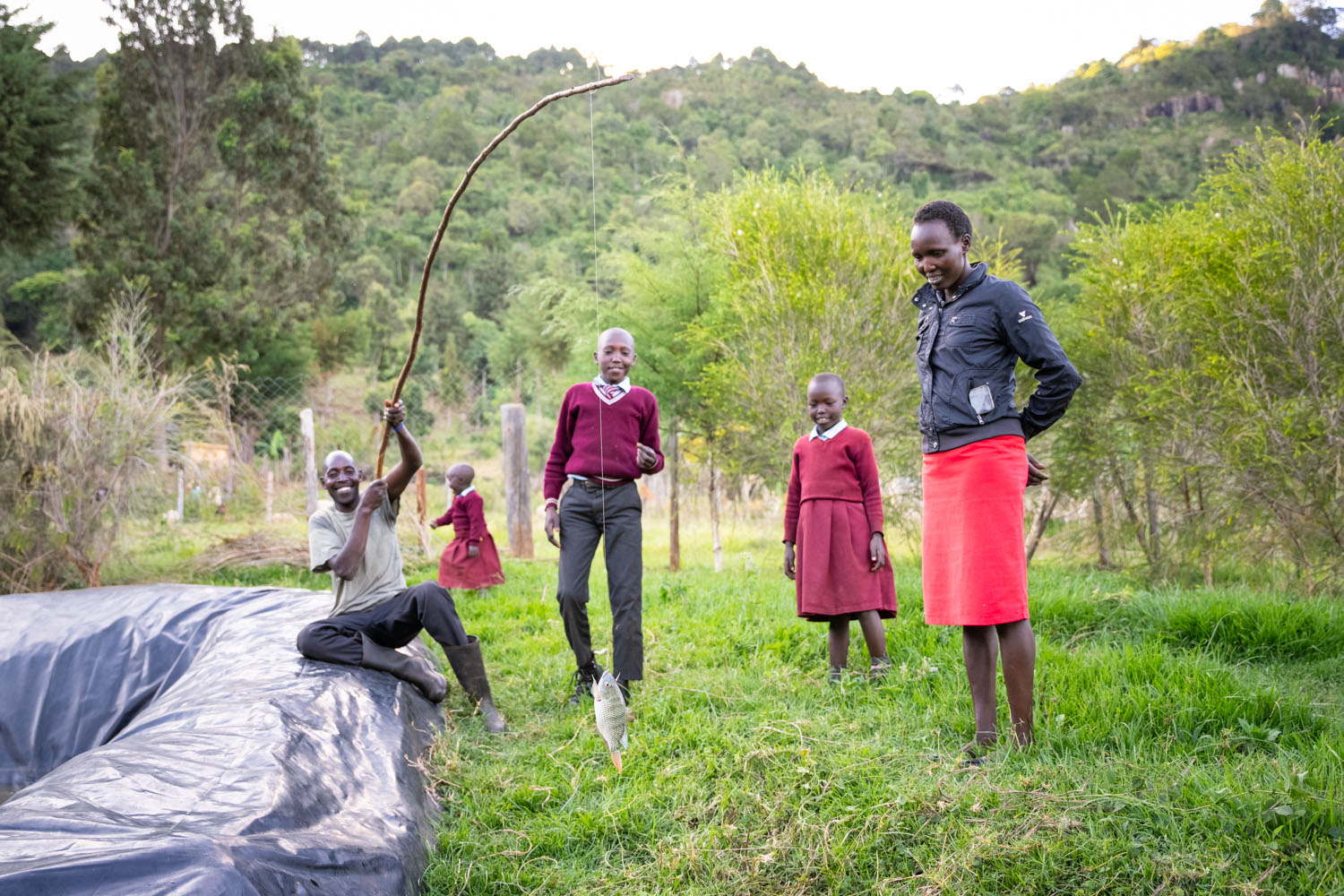 A Kenyan family of five stand beside their fishing pond, with the father proudly holding a caught fish on a fishing rod. 