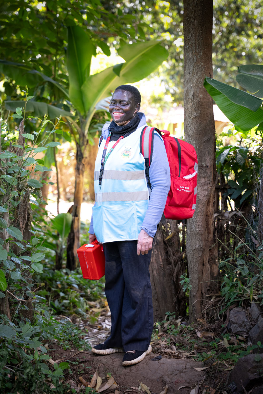 A Kenyan medical staff stands, smiling at the camera, carrying  a red backpack and a red first aid kit. 