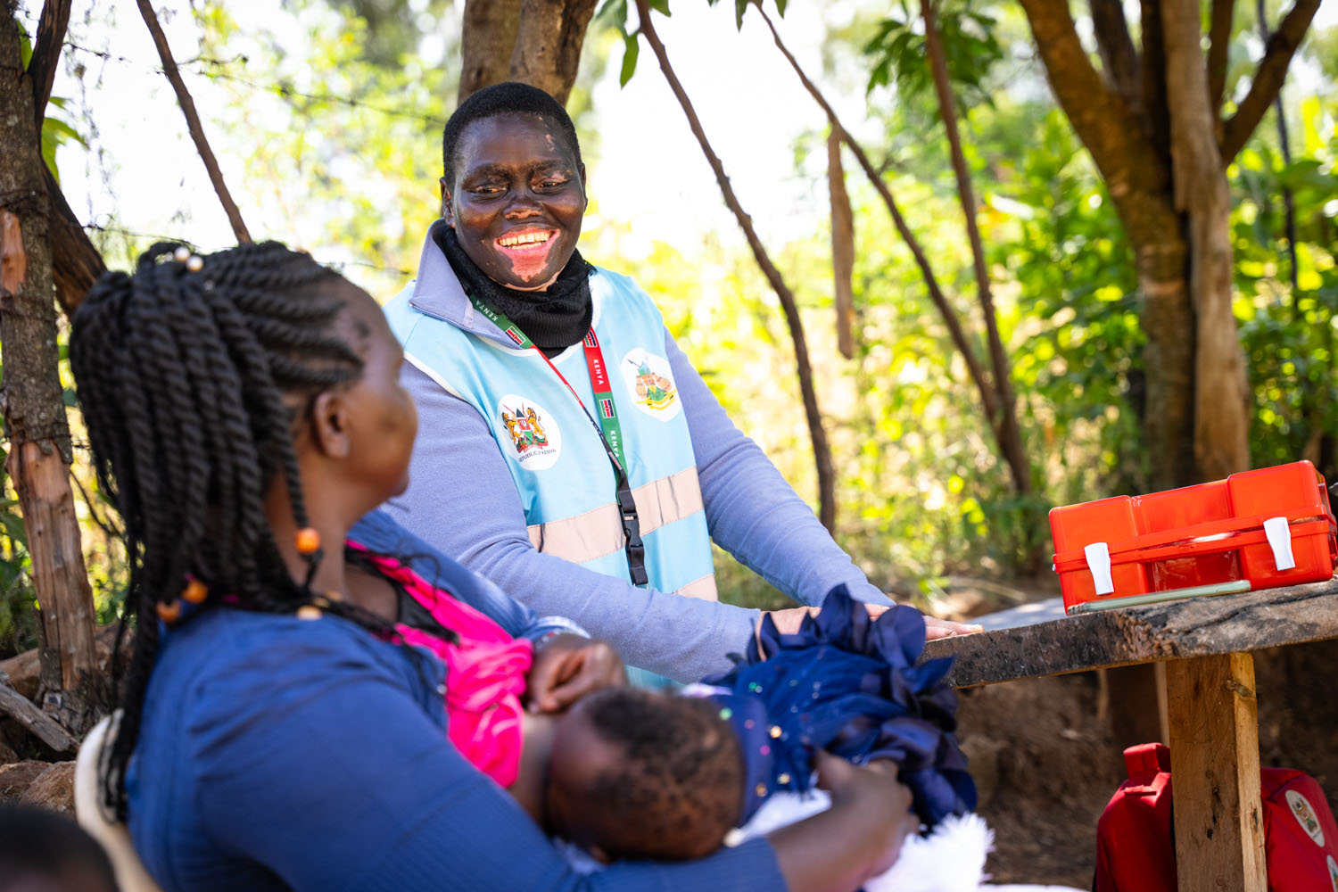 A Kenyan medical staff member talks with a mother and her child, with a first aid kit on the table. 