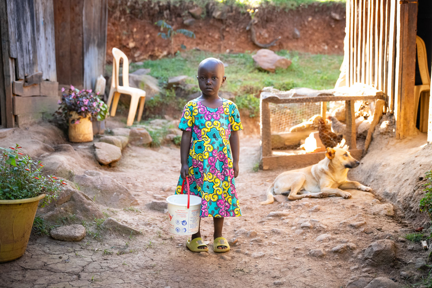 A young Kenyan girl stands in her front yard, holding a bucket. 
