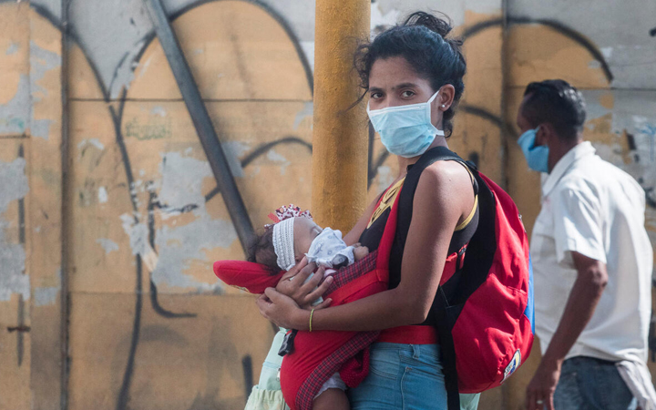 In Venezuela, a young woman with a baby past a wall of graffiti. She is wearing a mask for COVID-19 protection.
