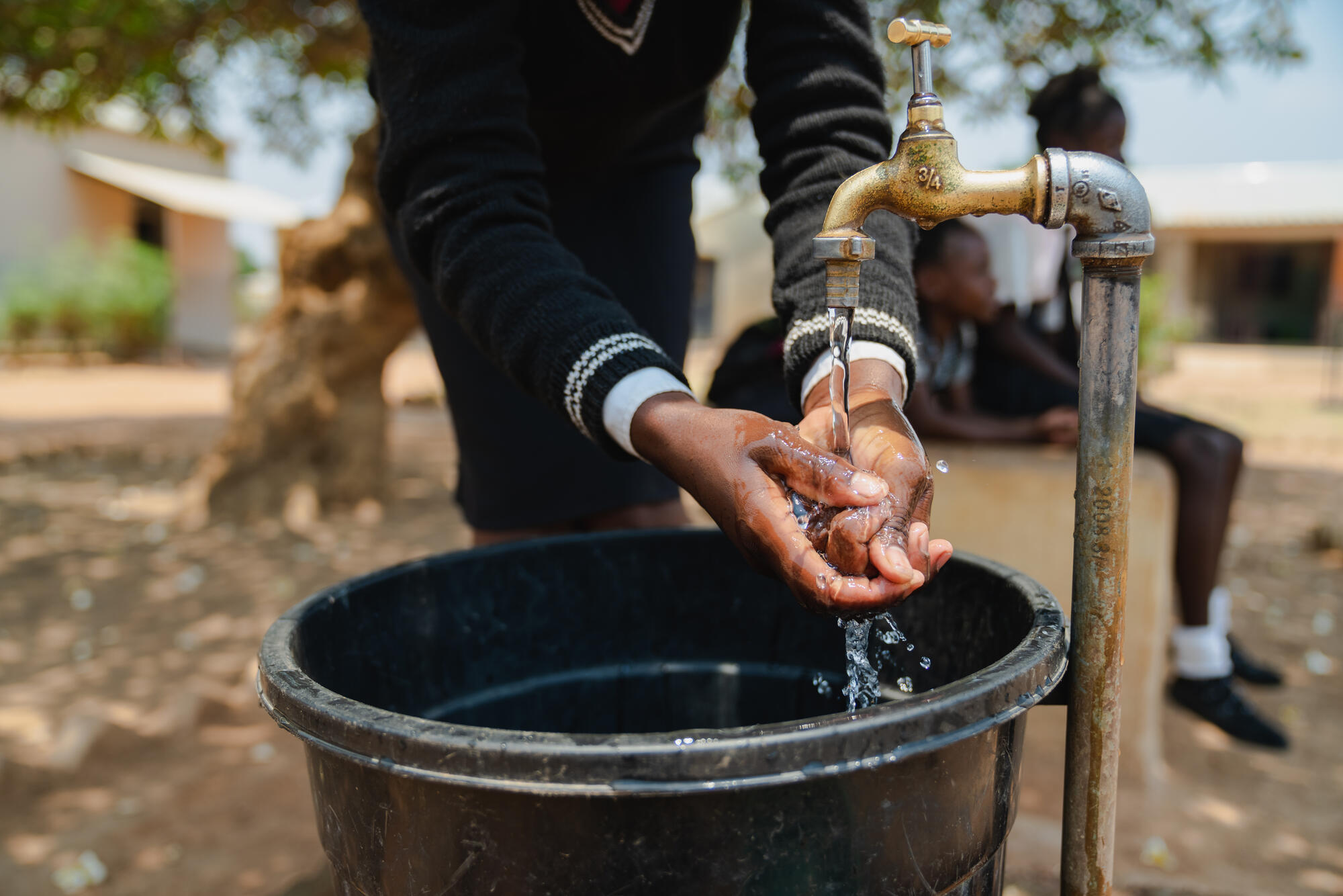 A person washes their hands at a clean water access point over a bucket.