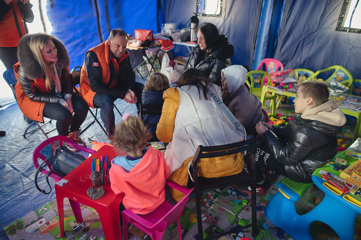 A group of children and adults sit talking in an area that has children’s tables, chairs and craft supplies.