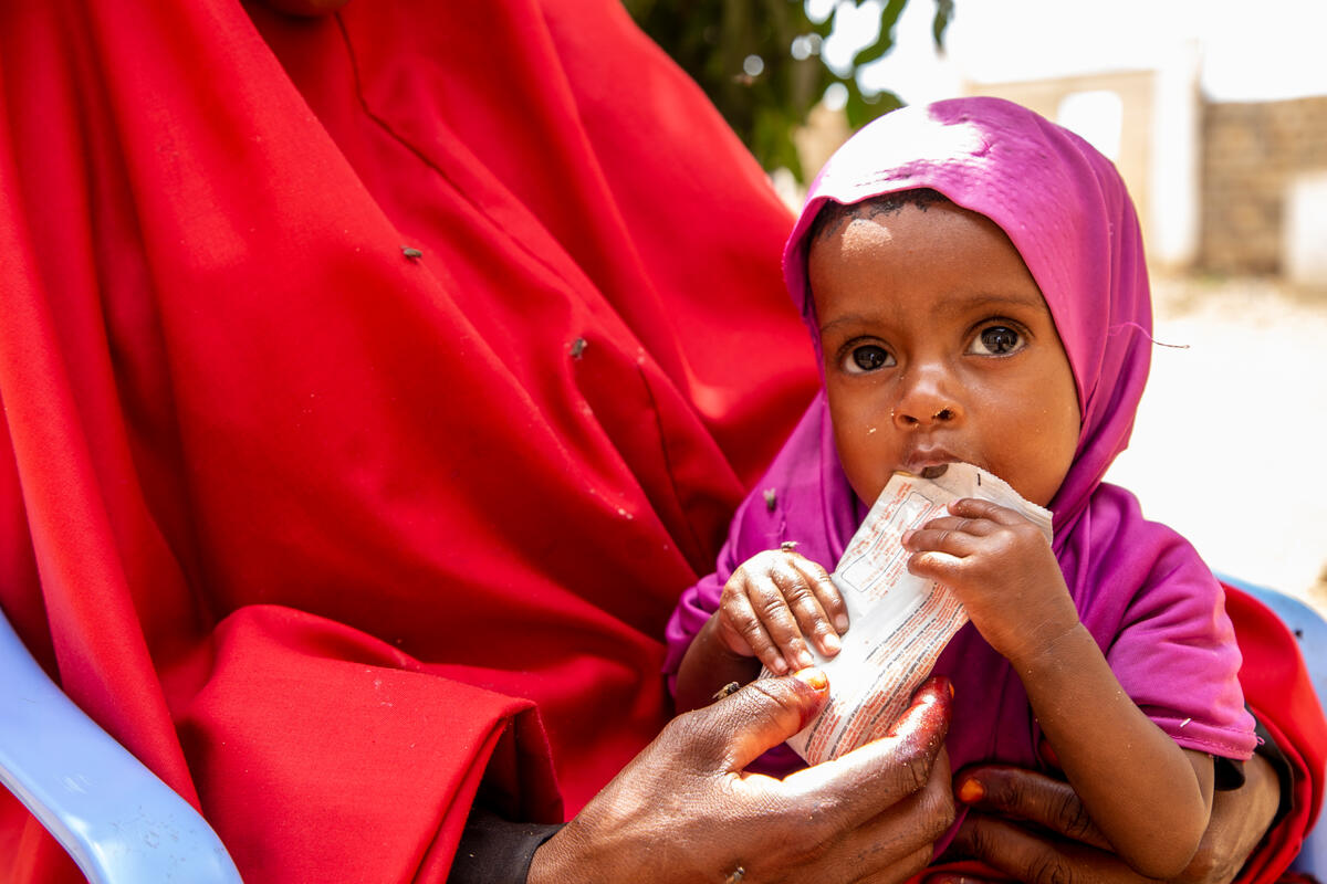 An infant sits in an adult’s arms, eating from a packet of food. 