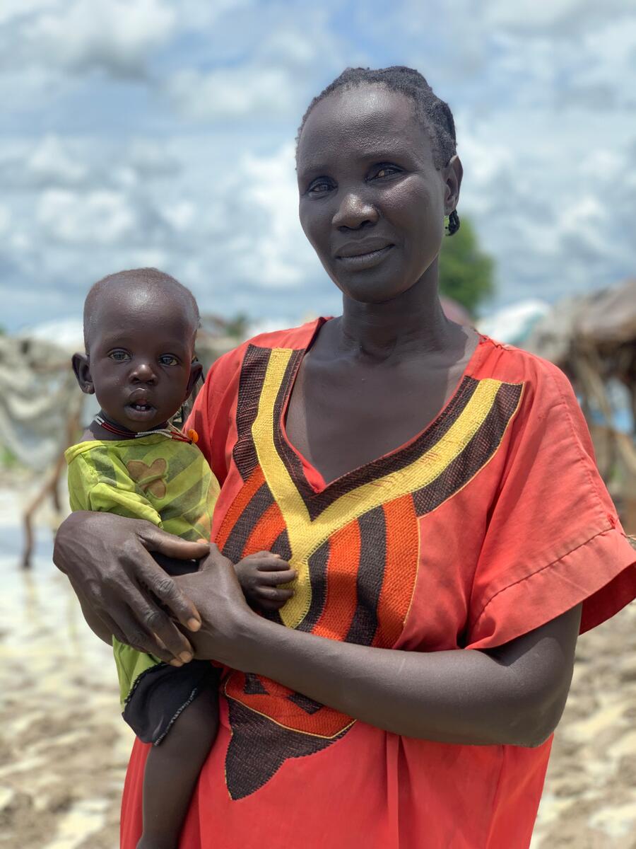 A mother wearing a red tunic stands with her baby on her hip.