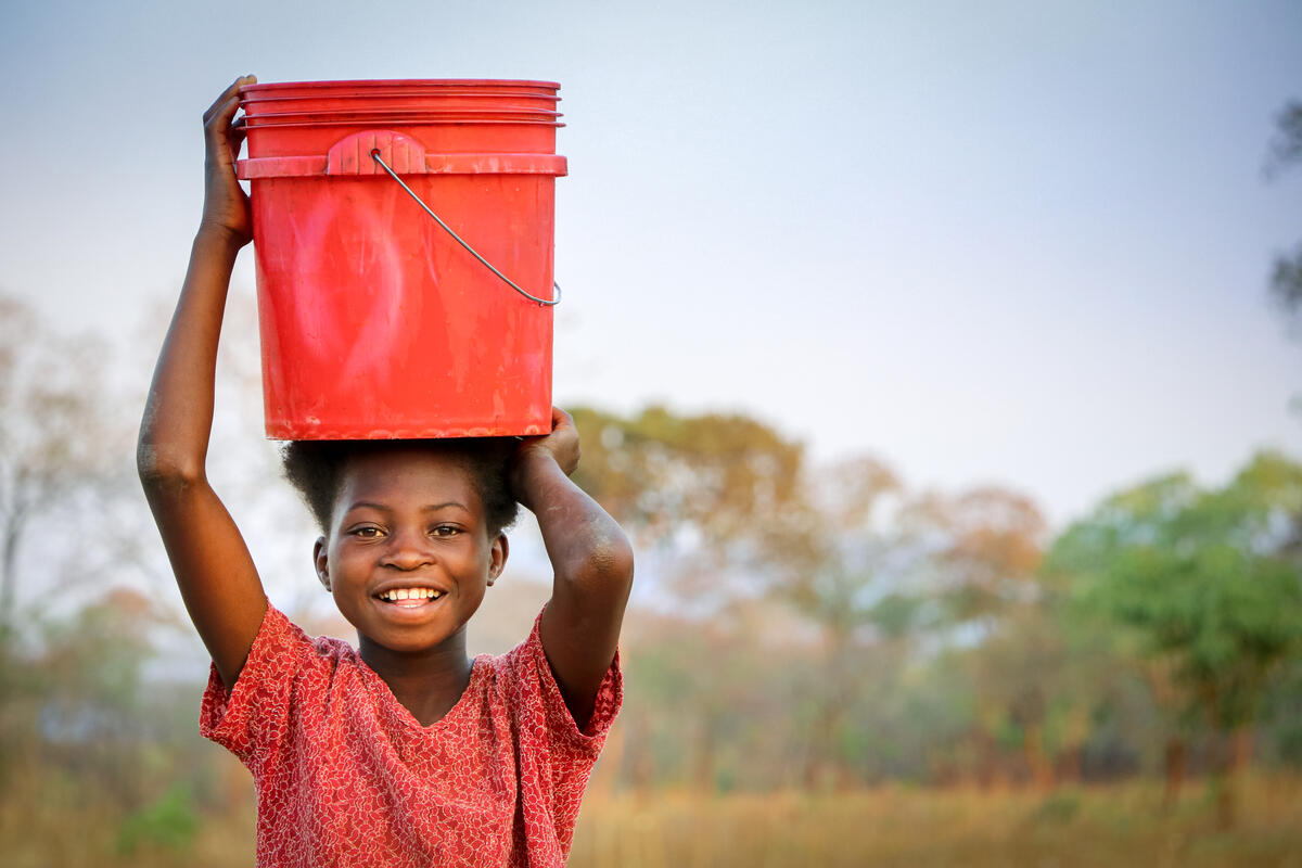 A child smiling as she holds a bucket over her head. 