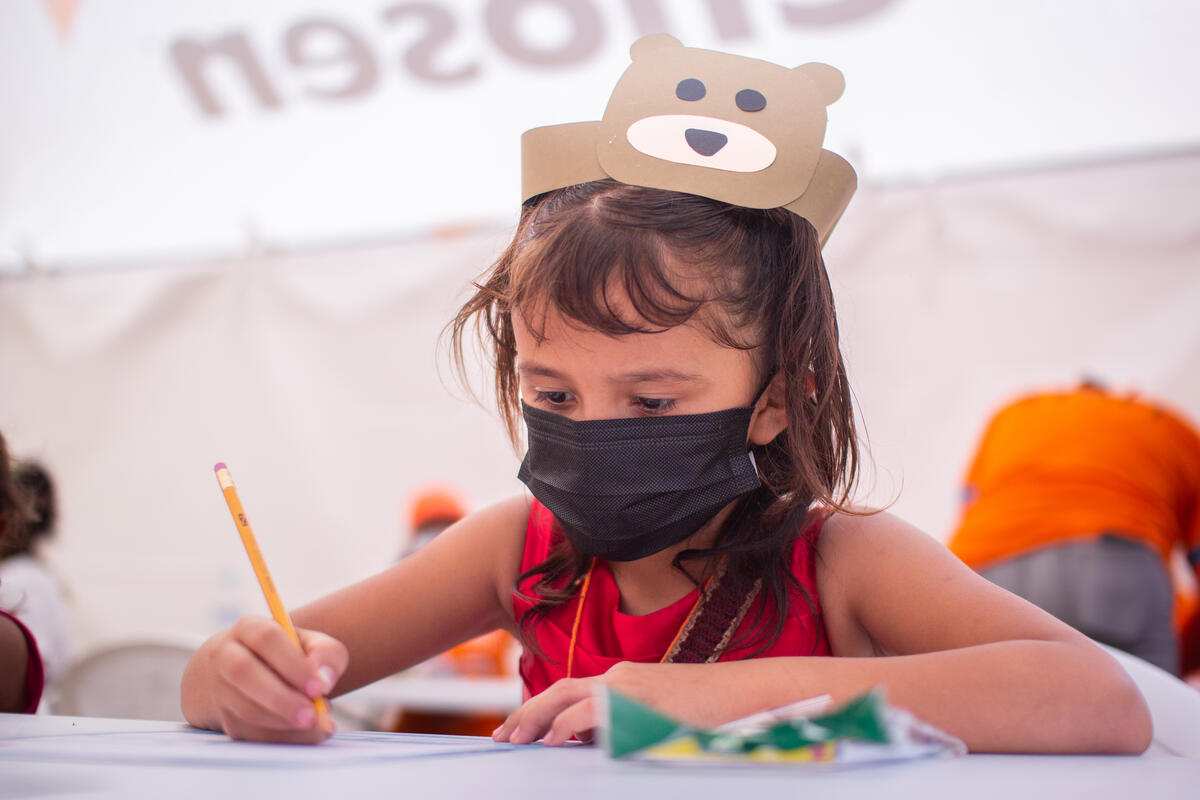 A child with a mask on writes on a paper. 