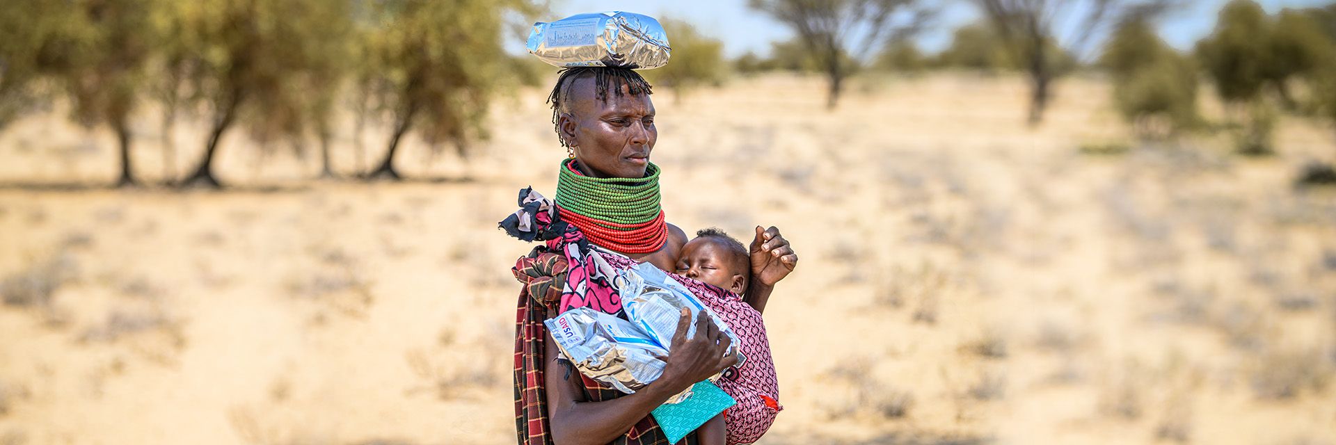 A woman outside wearing traditional garments holds her baby close to her chest in a cloth strap. 