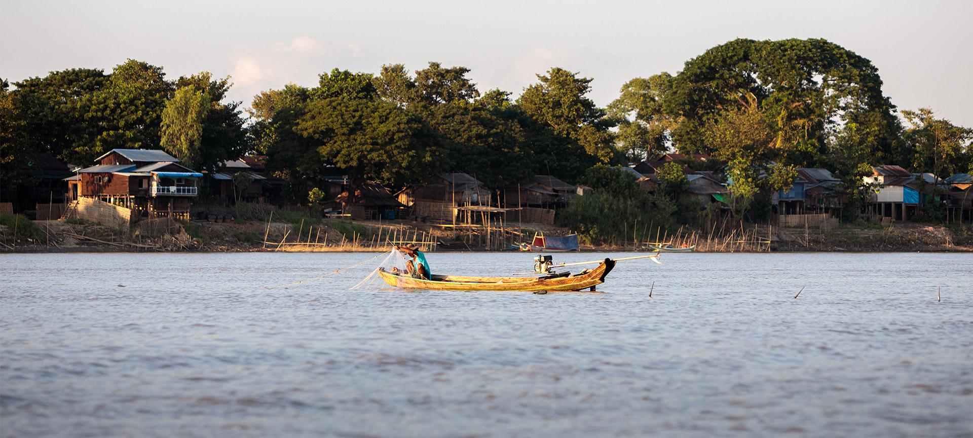 A photograph of a lake with forestry and small houses in the background along the shore. On the water, there is a fisherman in a small yellow canoe.