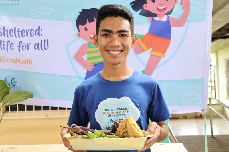 A teenaged Filipino boy wears a blue t-shirt and holds a bowl of vegetables.