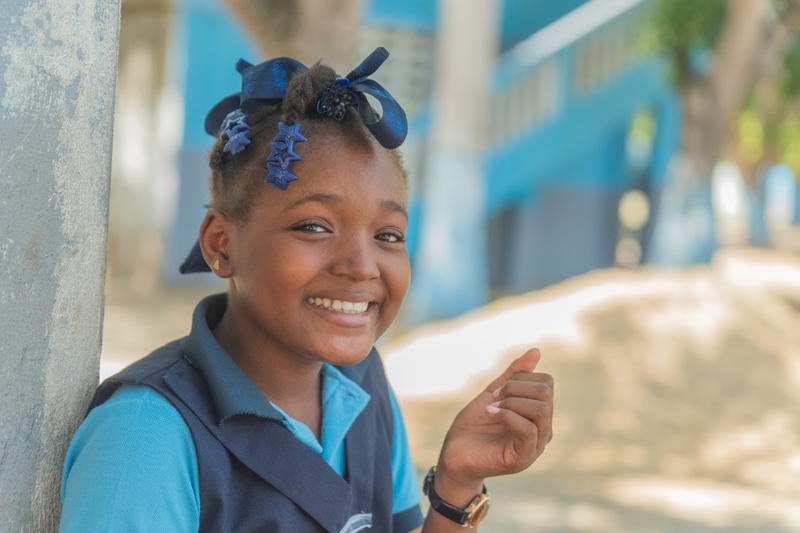 A Haitian girl wearing a blue school uniform smiles at the camera.