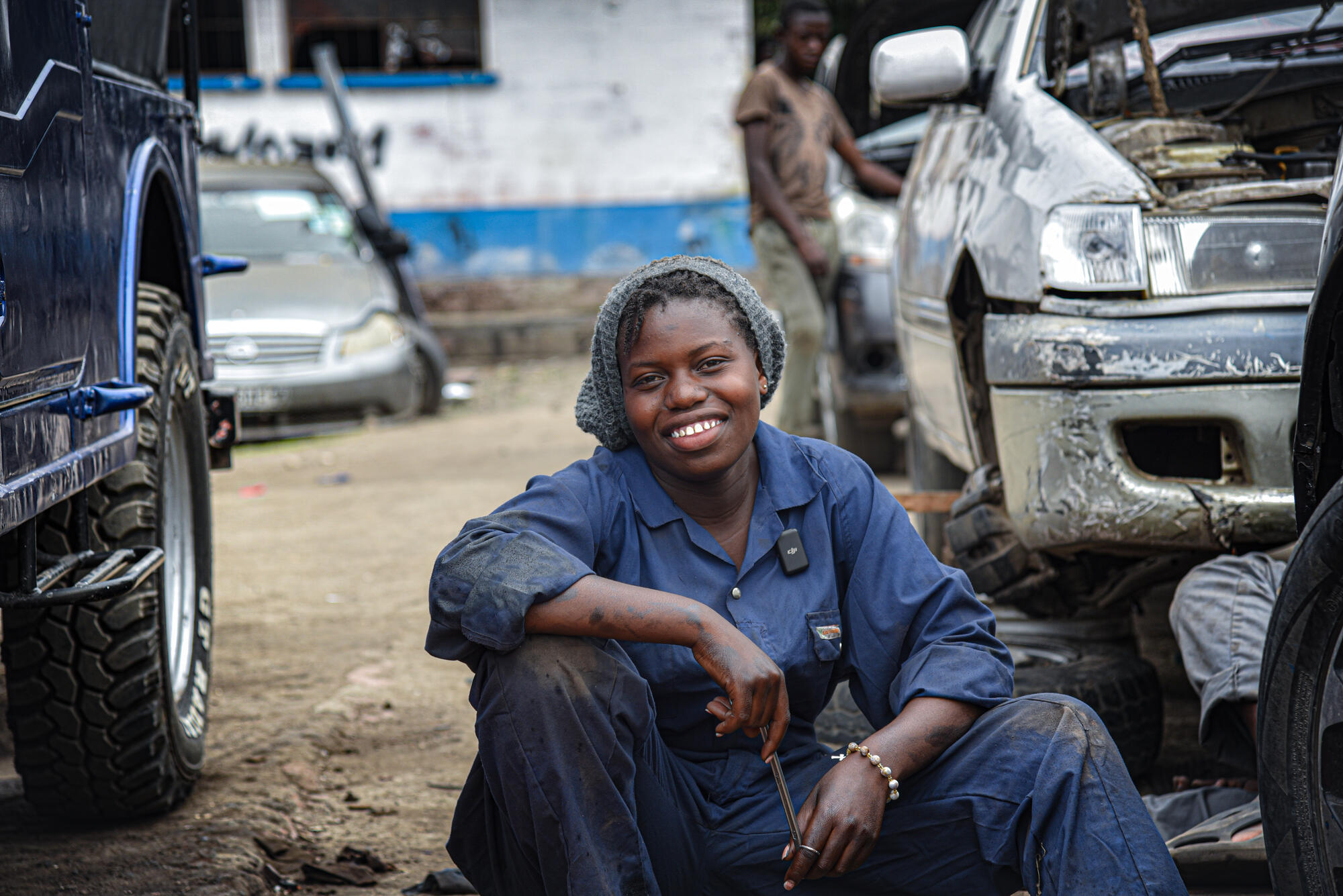 In Goma, DRC, where World Vision’s Raw Hope program is being implemented, a smiling young woman in navy blue coveralls holds a wrench, sitting on the ground between two vehicles.