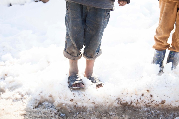 We see the feet of two children standing in ankle-deep snow, one child wearing sandals.