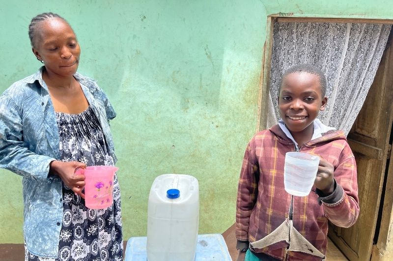 A young boy in Menkao, DRC, smiles while holding a clear cup of water; a woman stands beside him and a water jug sits on the table between them.