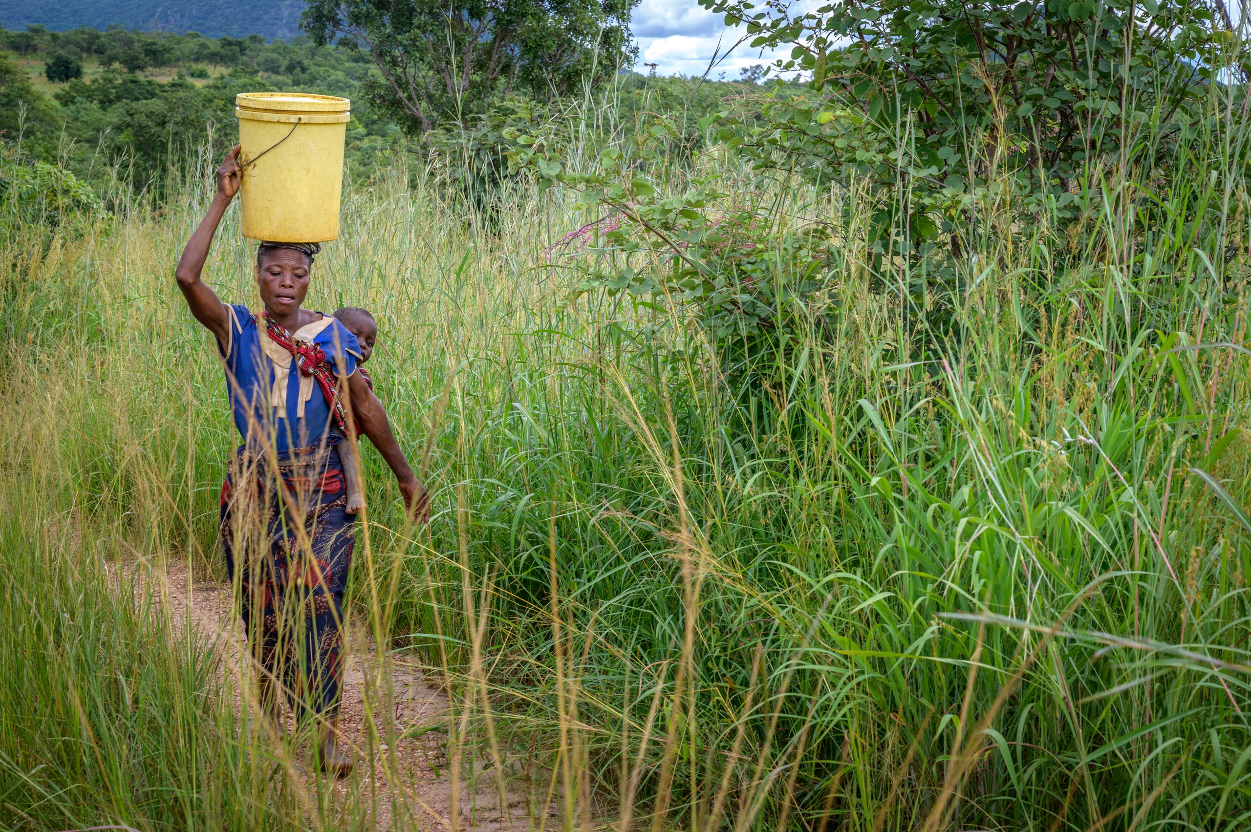 A woman carries her baby on her back and a yellow bucket of water on her head. 
