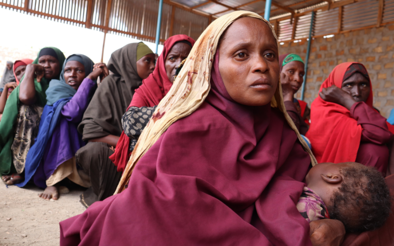 A group of women sit on the ground in a line, waiting.