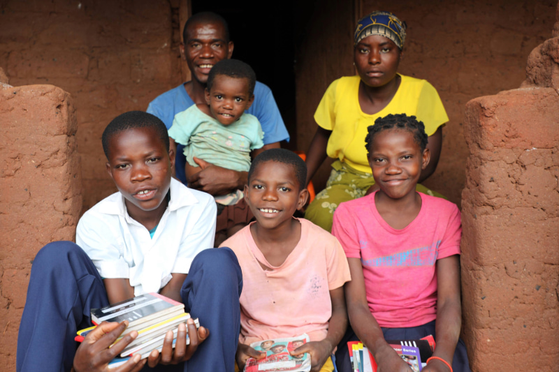 Family with children holding books outside a mud-brick home in a rural setting.  