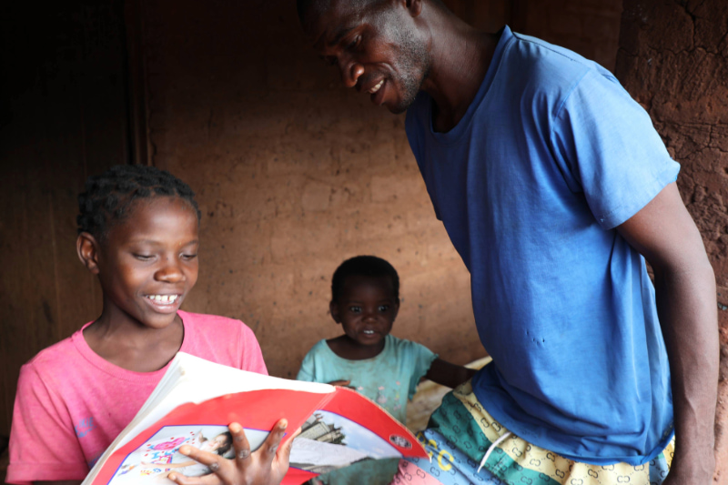 Daughter holds a book while showing her father her schoolwork.  