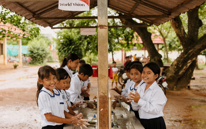 Students dressed in uniforms stand outside to wash their hands at a World Vision Water, Sanitation and Hygiene station outside of their school.