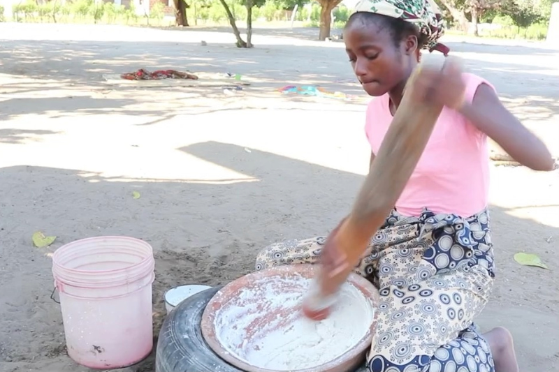 Young girl grinding grain outdoors with a wooden pestle.