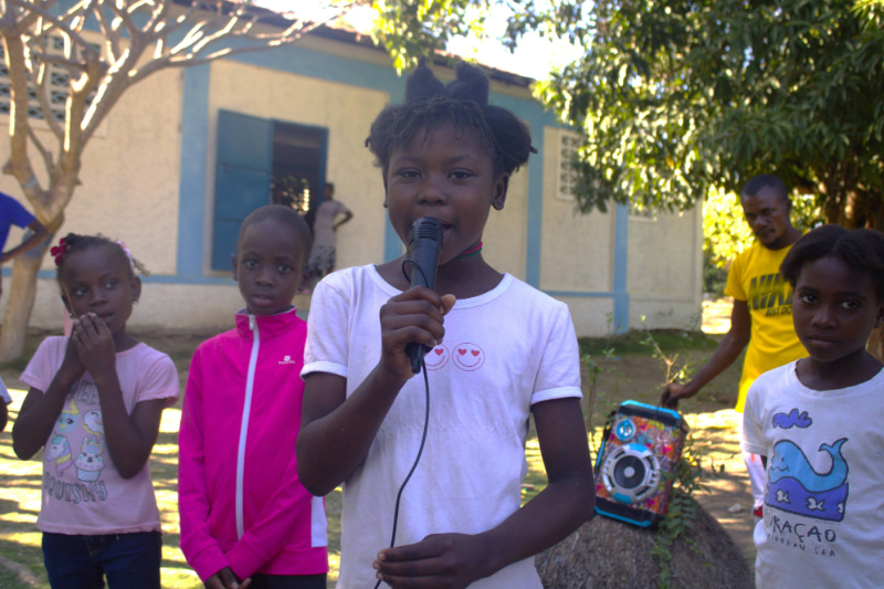 Young girl speaking into a microphone while other children stand around her.